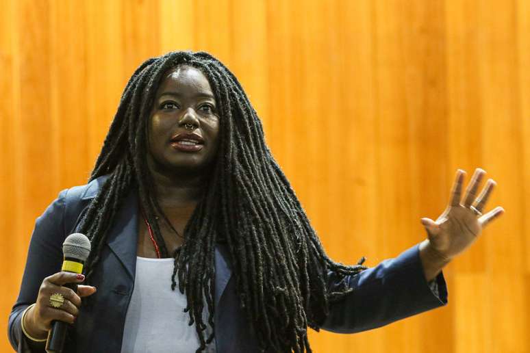 Imagem mostra a militante, pesquisadora e escritora Carla Akotirene. Ela é uma mulher negra retinta, com cabelo trançado.