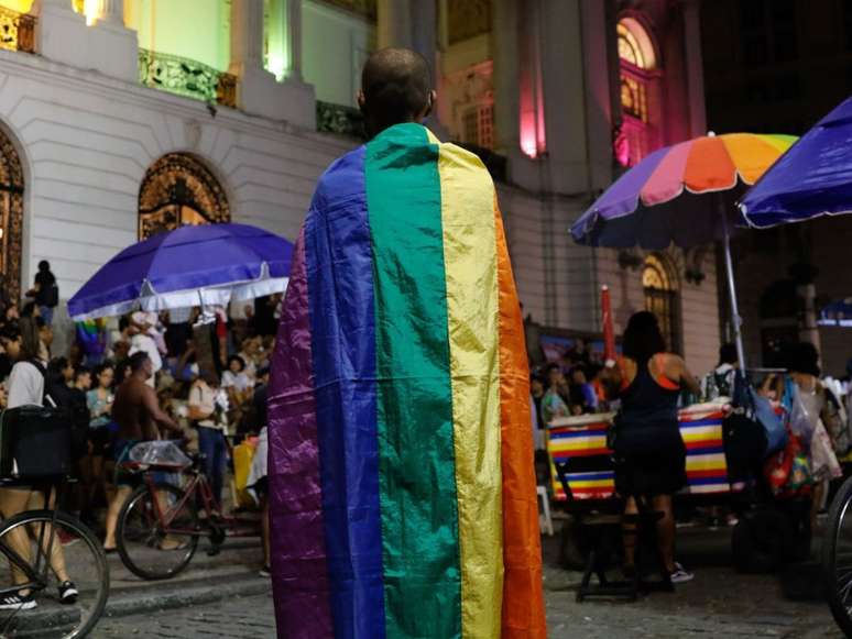 A imagem mostra um homem negro de costa, coberto com a bandeira da população LGBTQIA+