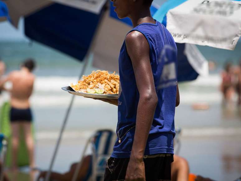 Imagem mostra um adolescente negro na praia de Guarujá levando uma porção de batata frita aos banhistas.