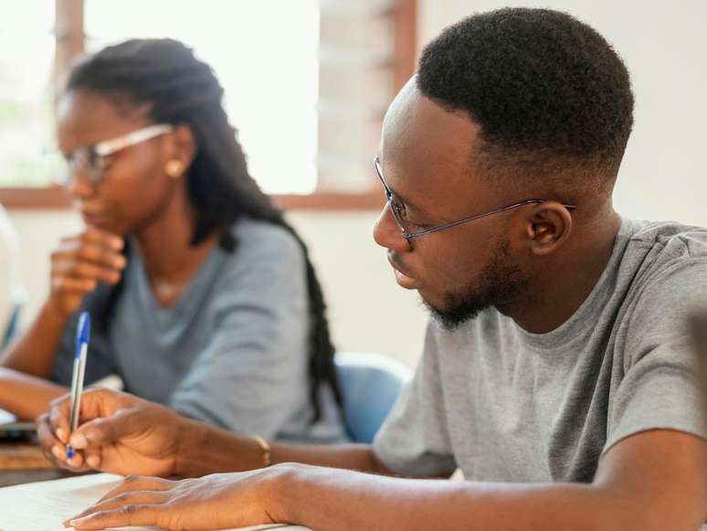 A imagem mostra um homem negro estudando, em destaque, e uma mulher negra atrás desfocada. Pessoas autodeclaradas negras serão contempladas por reserva de vagas no Concurso Nacional Unificado