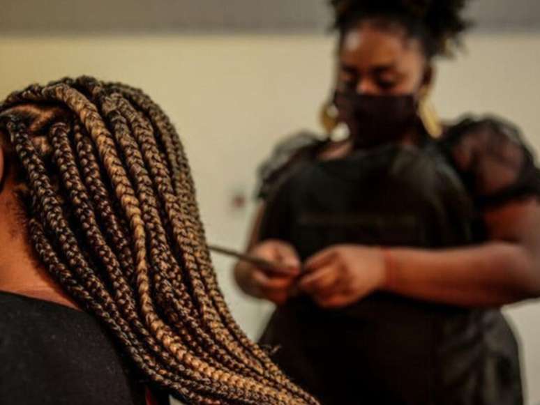 A imagem mostra uma mulher negra retinta fazendo tranças no cabelo de outra mulher.