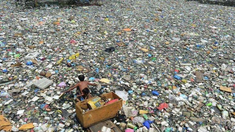 Pai e filho em um barco improvisado remam em rio cheio de lixo enquanto coletam garrafas plásticas que podem vender como recicláveis; a foto é de 2015 e foi registrada em Manila, Filipinas