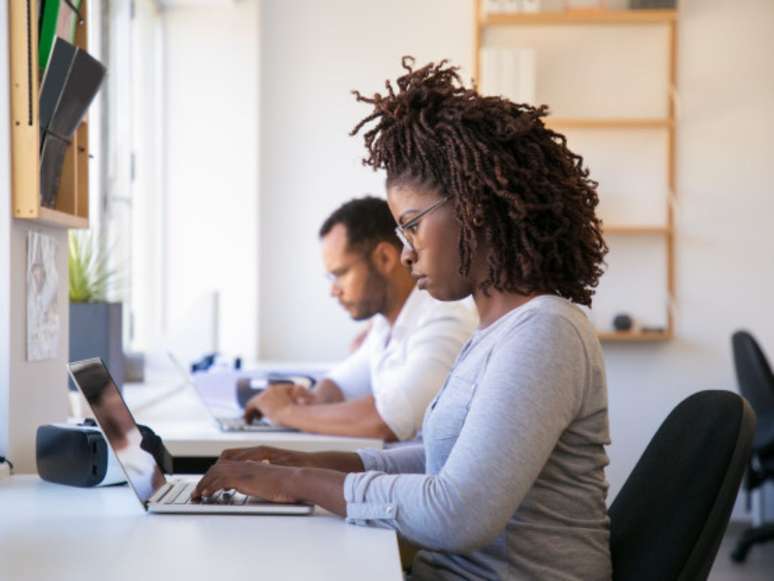 Imagem mostra uma mulher negra sentada em uma mesa digitando no computador, ao seu lado, em segundo plano, está um homem negro realizando a mesma atividade.