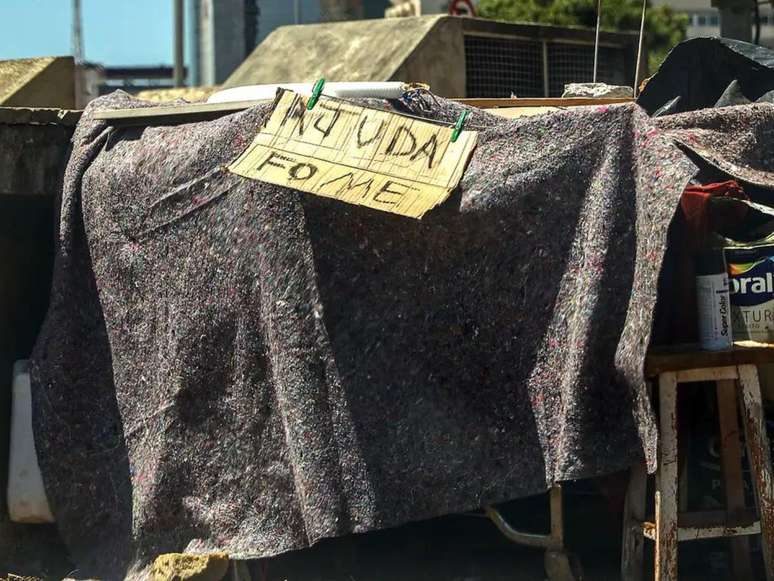 Imagem mostra o acampamento de pessoas em situação de rua, no qual há uma placa com os dizeres: "ajuda, fome".