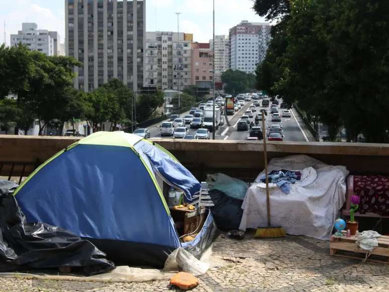 Imagem mostra um acampamento de pessoas em situação de rua, em contraste, prédios aparecem em segundo plano para ressaltar a desigualdade presente no Brasil.