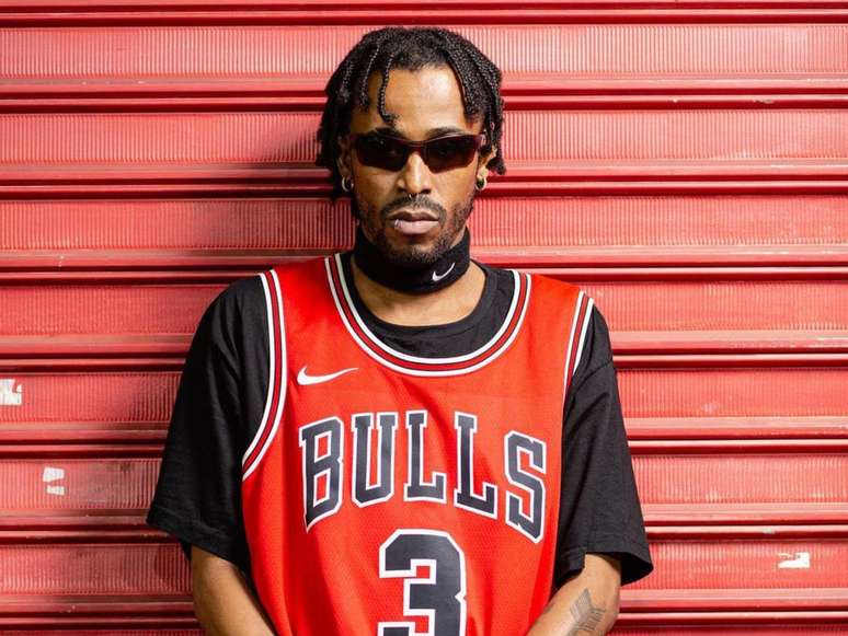 A imagem mostra o compositor e cantor de rap Nego Max em frente a uma porta de aço vermelha.