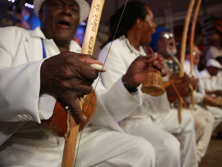A imagem mostra um homem negro em destaque, junto de outros 5 homens desfocados, tocando instrumentos. Todos são capoeiristas, público do evento que acontece em Salvador, entre os dias 24 e 27 de janeiro.