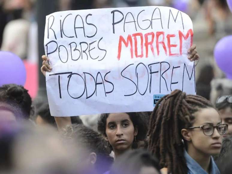 A imagem mostra uma mulher segurando um cartaz com os dizeres “ricas pagam, pobres morrem e todas sofrem” em protesto.