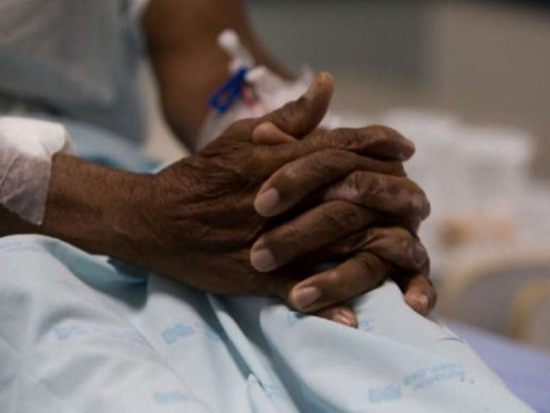 A imagem mostra as mãos de um paciente negro com acesso nos antebraços para medicação intravenosa.