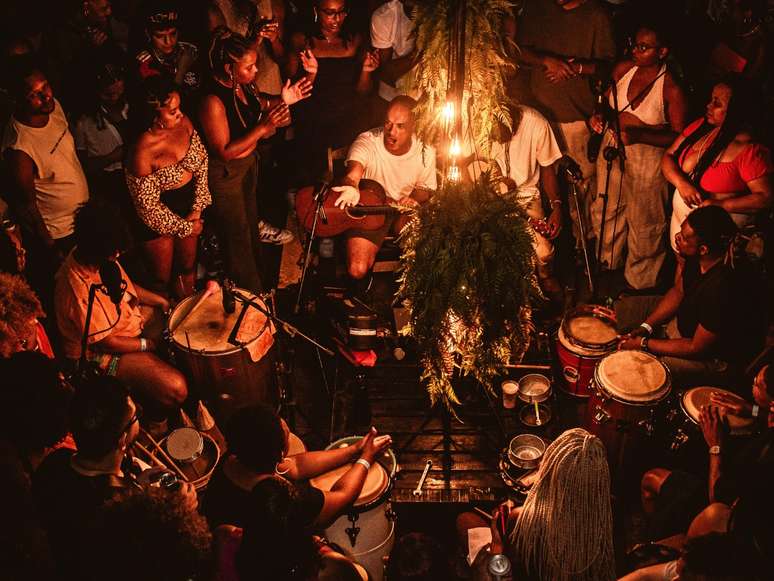 A imagem mostra pessoas a roda de samba "Poeira Pura", tocando no centro da imagem. Ao redor, o p&uacute;blico do evento contempla o show.
