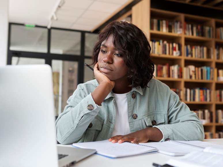 Imagem mostra uma mulher negra, sentada à mesa e olhando para um notebook.