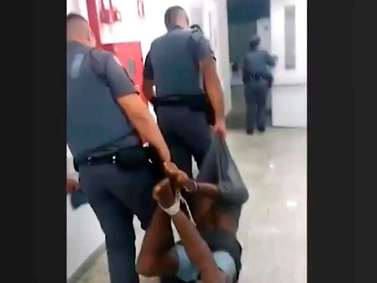 A imagem mostra dois policiais militares carregando um homem negro amarrado pelos pés e mãos.