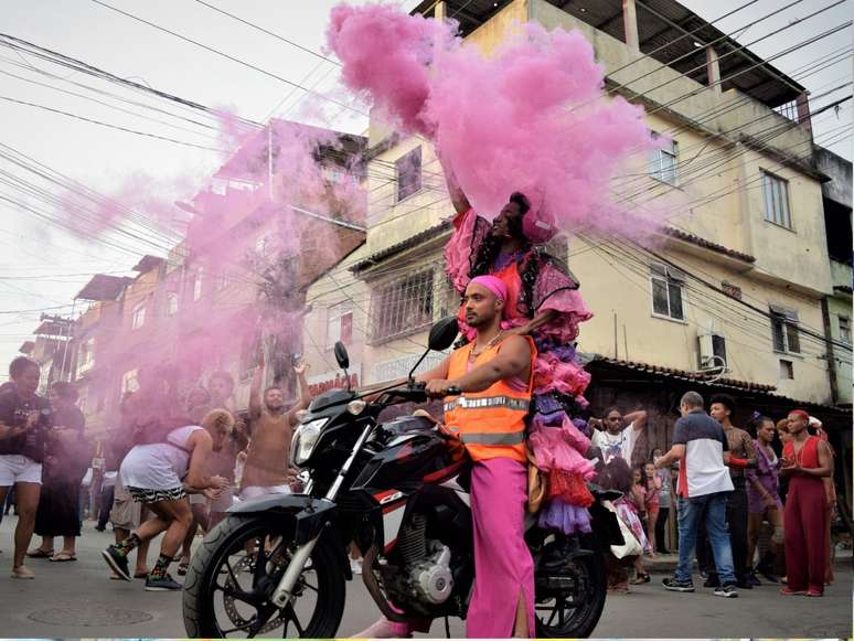 Imagem mostra a performance de Milu Almeida e Luiz Otávio na favela da Nova Holanda (RJ). Na foto, os artistas aparecem em cima de uma moto com trajes na cor rosa e laranja, enquanto dezenas de pessoas os cercam.