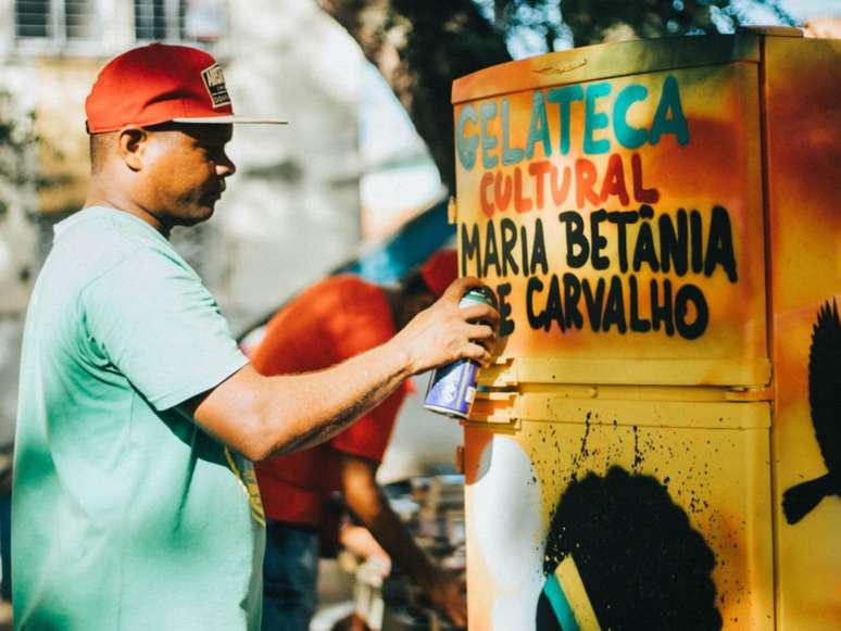 A imagem mostra um homem negro com uma lata de tinta em frente a uma geladeira com os dizeres “Gelateca Cultural Maria Betânia de Carvalho”.