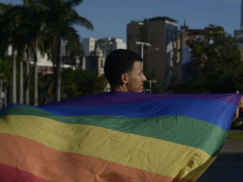 A imagem mostra uma pessoa negra pertencente à população LGBTQIAPN+ carregando a bandeira da sigla.