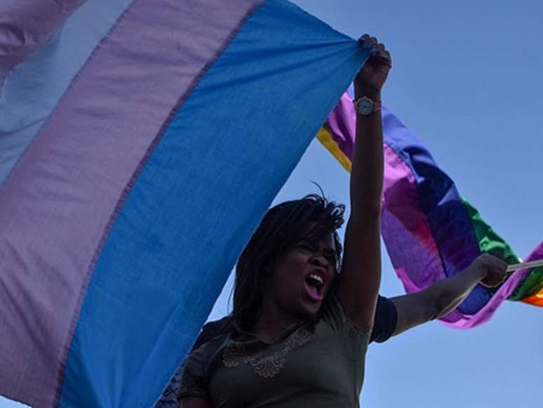 A imagem mostra uma pessoa negra segurando uma bandeira da população trans. Relatório revela que pessoas afetadas pelo racismo são maioria nos registros de assassinatos.