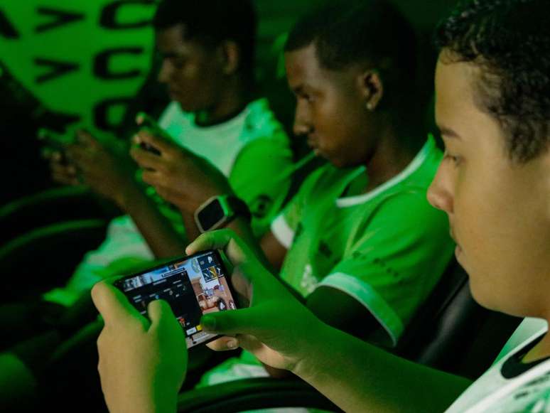 A imagem mostra três jovens negros, sentados em cadeiras games com o celulares das mãos jogando uma das modalidades da Copa AFG.