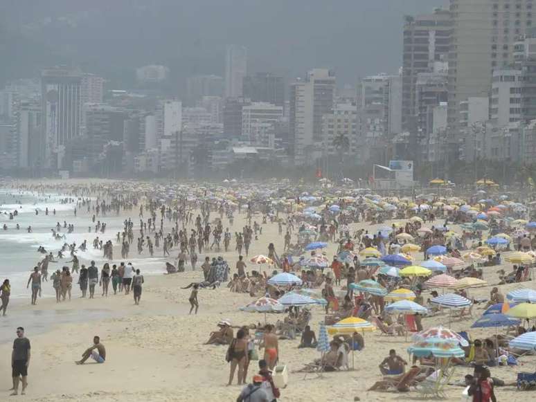 A imagem mostra uma praia do Rio de Janeiro cheia de banhistas na área e na areia.