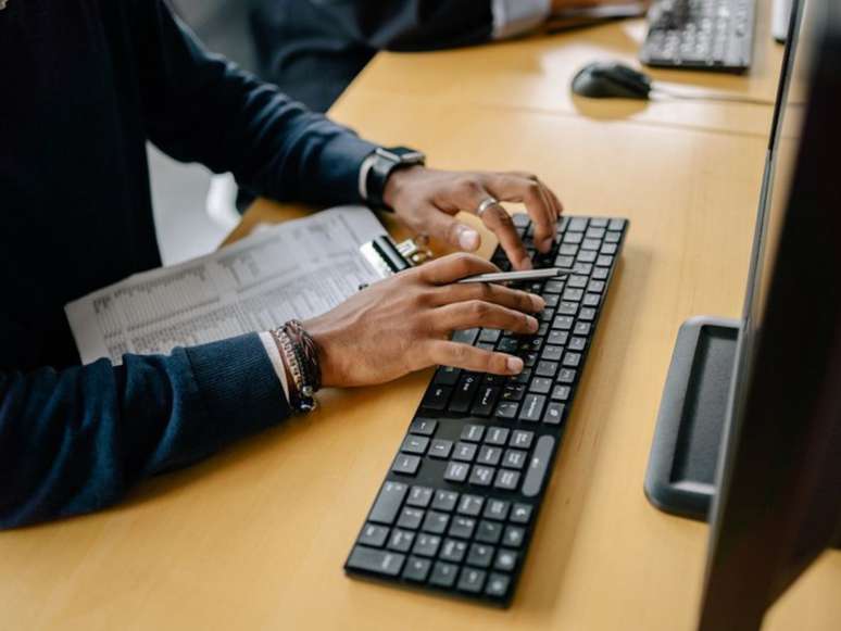 A imagem mostra as mãos de um homem negro, um teclado e o monitor de computados, além de uma prancheta.