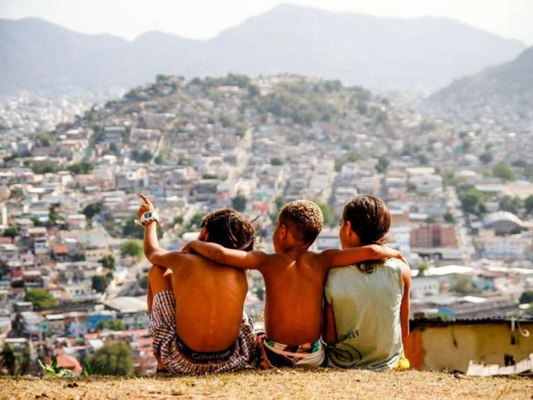 Imagem mostra três garotos negros de costas para a câmera. Ao fundo, aparece uma favela do Rio de Janeiro.