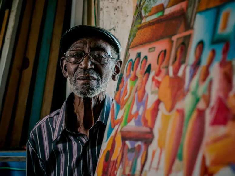 A imagem mostra o pintor e escultor João Cândido, um homem negro, idoso ao lado de uma de suas telas.