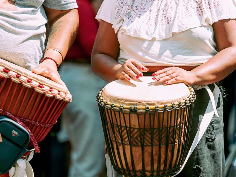 Imagem em plano fechado mostra as mãos de uma mulher tocando um tambor africano.