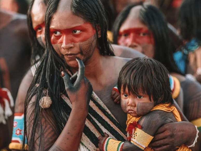 Imagem mostra uma mãe de ascendência indígena segurando seu filho pequeno em seu colo.