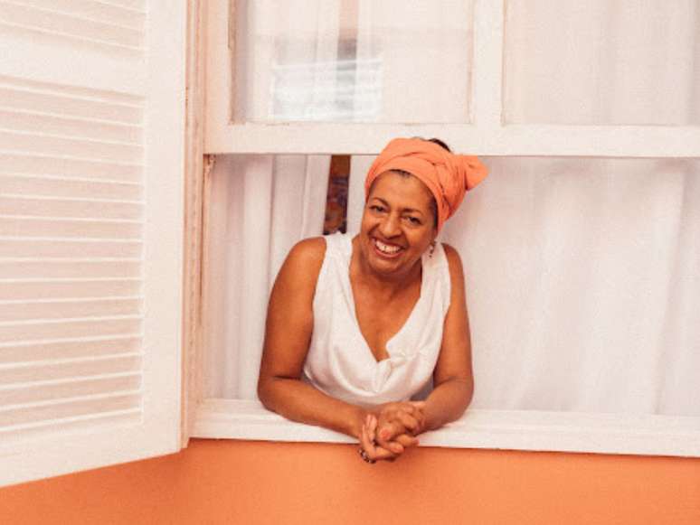 Na foto, Maria Helena, embaixatriz do samba paulista, posa sorridente em uma janela.