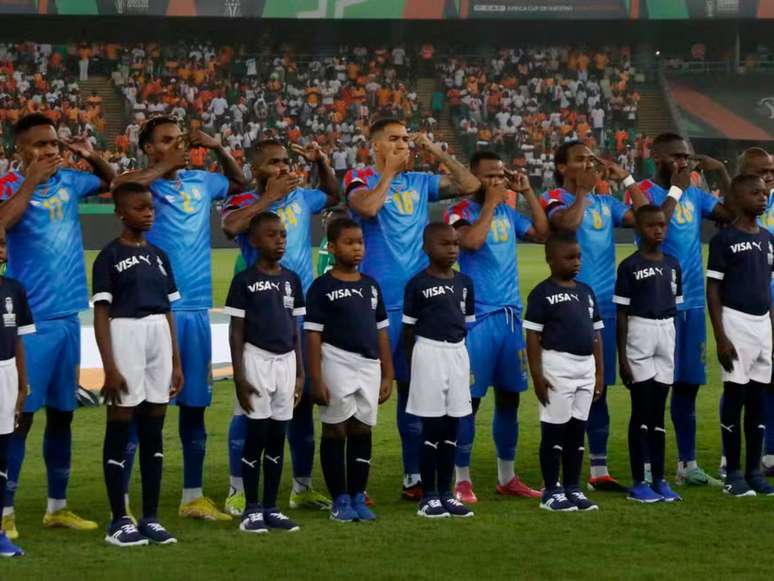 A imagem mostra jogadores de futebol em campo pela Copa Africana de Nações protestando contra o genocídio no país. Os atletas estão com uma mão na boca e a outra apontada para a cabeça.