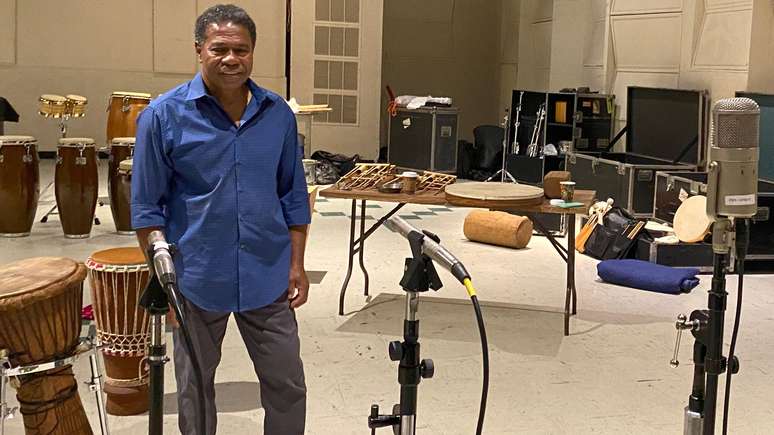 Paulinho da Costa, de camisa azul e em pé, em estúdio de gravação com microfones e instrumentos musicais