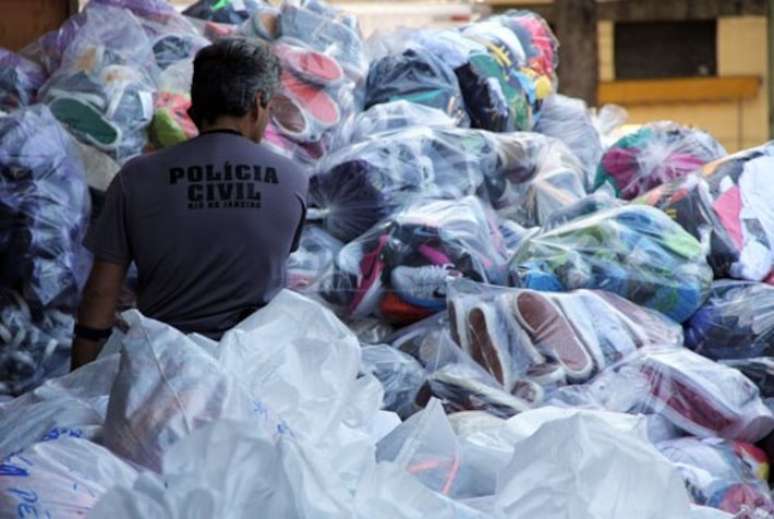 Agente da Polícia Civil em meio a uma pilha de produtos apreendidos durante operação contra pirataria no Rio de Janeiro.