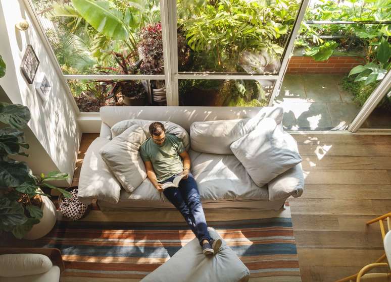Mezanino é transformado em quarto nesta cobertura de 95 m². Projeto de DCC Arquitetura. Na foto, sala de estar vista de cima, tapete listrado, sofá cinza, grandes janelas com vista para árvores.