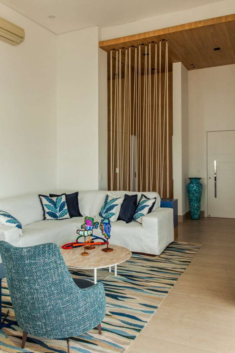 Apê tem décor inspirado na praia com toques azuis e porcelanato areia. Projeto de Korman Arquitetos. Na foto, sala de estar com pé direito alto, sofá branco, tapete listrado, mesa de centro.