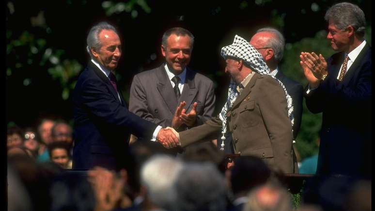 O líder da OLP, Yasser Arafat (à direita), apertando a mão do ministro das Relações Exteriores de Israel, Shimon Peres, na assinatura do Acordo de Oslo. O então presidente dos EUA, Bill Clinton (à direita), aparece batendo palmas