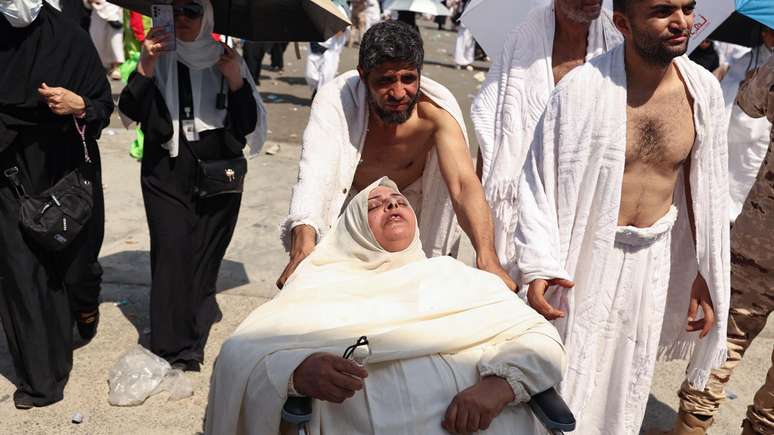 Uma mulher sofrendo com o calor escaldante é empurrada em uma cadeira de rodas, enquanto os peregrinos chegam para realizar o ritual simbólico de 'apedrejamento do diabo'