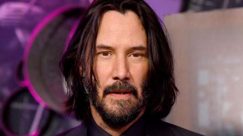 Imagem do rosto de Keanu Reeves, um homem branco, de barba, com o cabelo escuro e liso pouco acima do ombro  