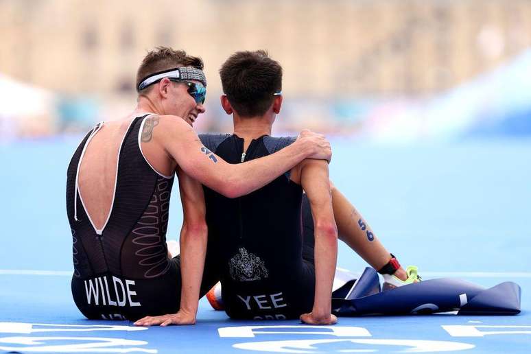 Hayden Wilde de Nueva Zelanda acaba de perder la medalla de oro en triatl&oacute;n masculino, pero eso no le impide animar a su amigo, el brit&aacute;nico Alex Yee, quien exhausto celebra en el suelo el oro conseguido.