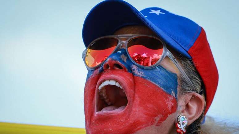 Manifestante com o rosto pintado de vermelho e azul, usando óculos e boné também nas cores azul e vermelho