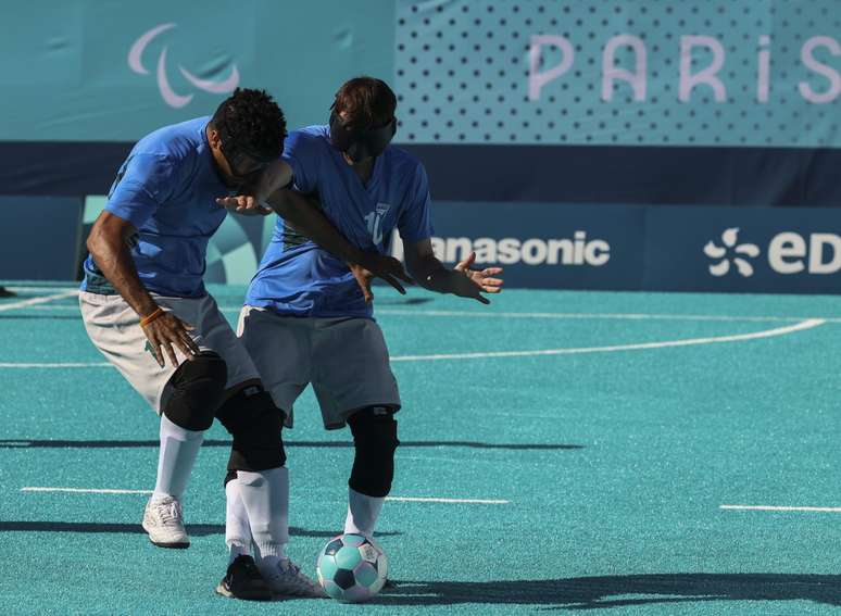 Ricardinho e Jardiel, ambos com uniforme de camisa azul, short branco e meião branco, disputam bola sobre a grama azul piscina do Estádio da Torre Eiffel, com placas de publicidade ao fundo 