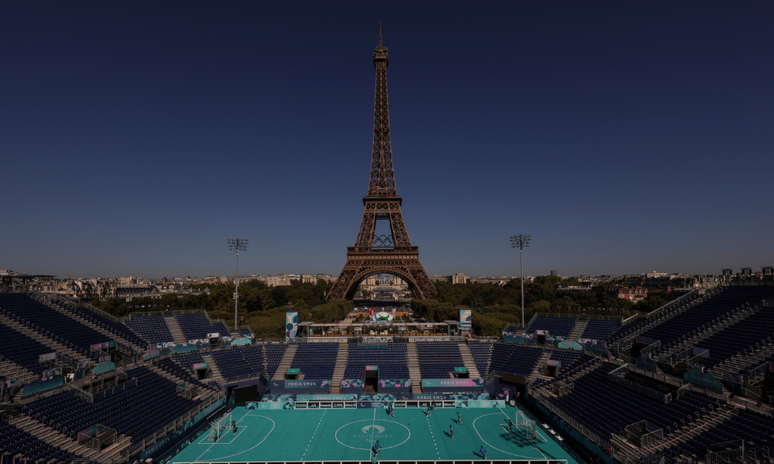 Imagem panorâmica do Estádio da Torre Eiffel, contando com o campo embaixo, cuja grama é azul piscina; as arquibancadas azuis marinho ao redor; e a torre ao fundo 