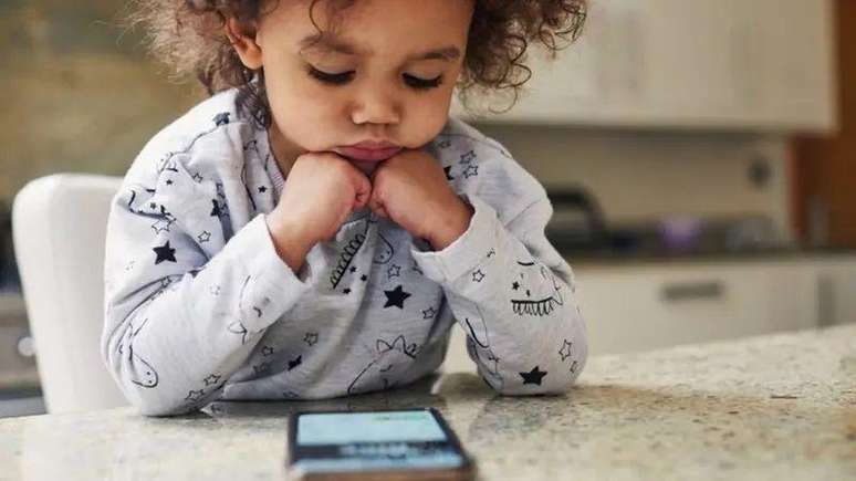 A foto mostra uma criança pequena, de cabelos cacheados e pele morena, sentada, com a cabeça apoiada nas mãos e olhando para um celular sobre a mesa. 