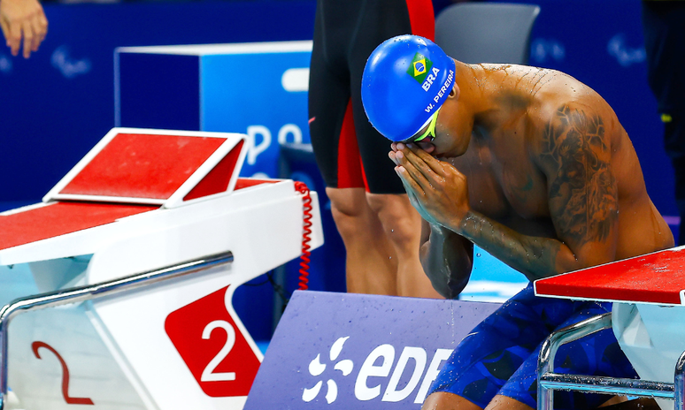 Wendell está com touca, sem camisa e com calção. Ele abaixa a cabeça, à beira da piscina, e junta as duas mãos ao rosto, em uma espécie de oração 