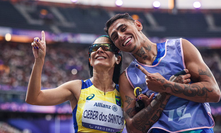 Jerusa Geber e Gabriel Garcia sorriem e fazem o número "1" com o dedo. Ele usa colete, enquanto ela tem uniforme amarelo e usa óculos de corrida 