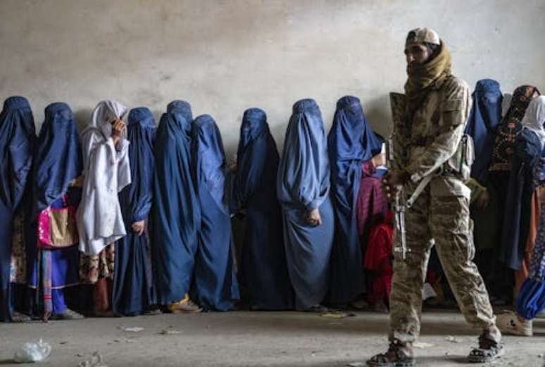 Um combatente do Talibã monta guarda enquanto mulheres esperam para receber rações de alimentos distribuídas por um grupo de ajuda humanitária em Cabul, Afeganistão, em 23 de maio de 2023. AP Photo/Ebrahim Noroozi