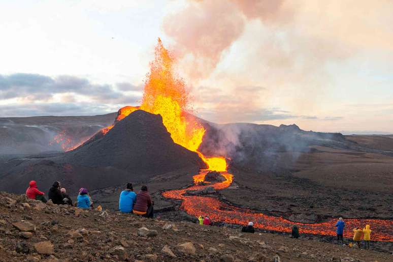 Um pequeno grupo de pessoas senta-se em um cume rochoso observando um vulcão ardente entrar em erupção à distância com fumaça e lava brilhante.