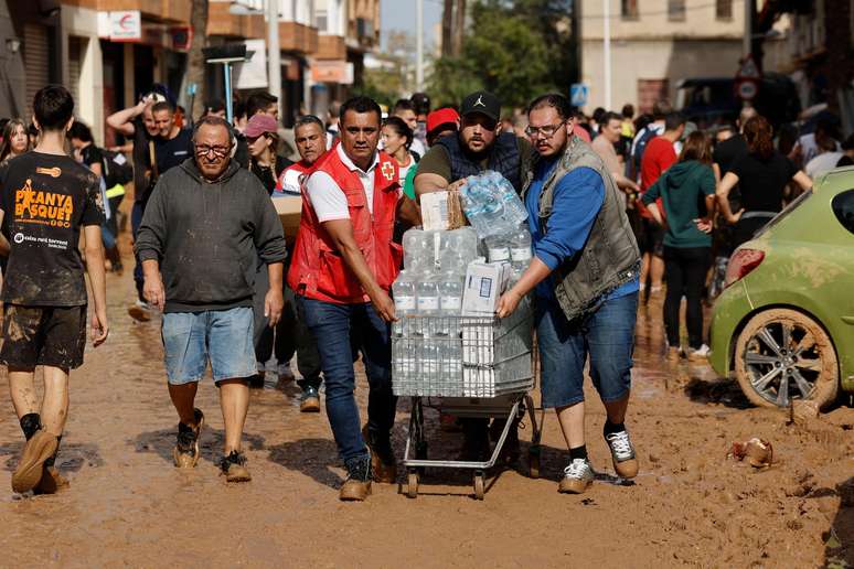 Voluntários levam garrafas d'água em carrinho de supermercado; eles caminham sobre rua repleta de lama