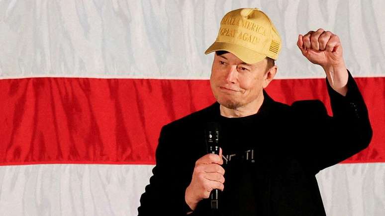 Elon Musk vestindo um blazer preto e um boné amarelo onde se lê "Make America Great Again", slogan da campanha de Trump, com a mão em punho e segurando um microfone na outra mão. 