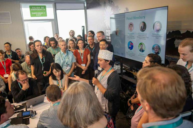 tela de computador, pessoas em p&eacute; ao redor ouvindo uma mulher com microfone