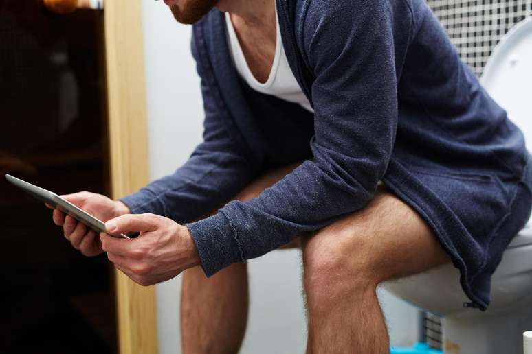 Homem sentado no vaso sanitário com um tablet nas mãos.