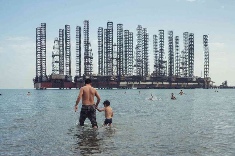 Um homem e seu filho na praia com refinarias de petróleo atrás